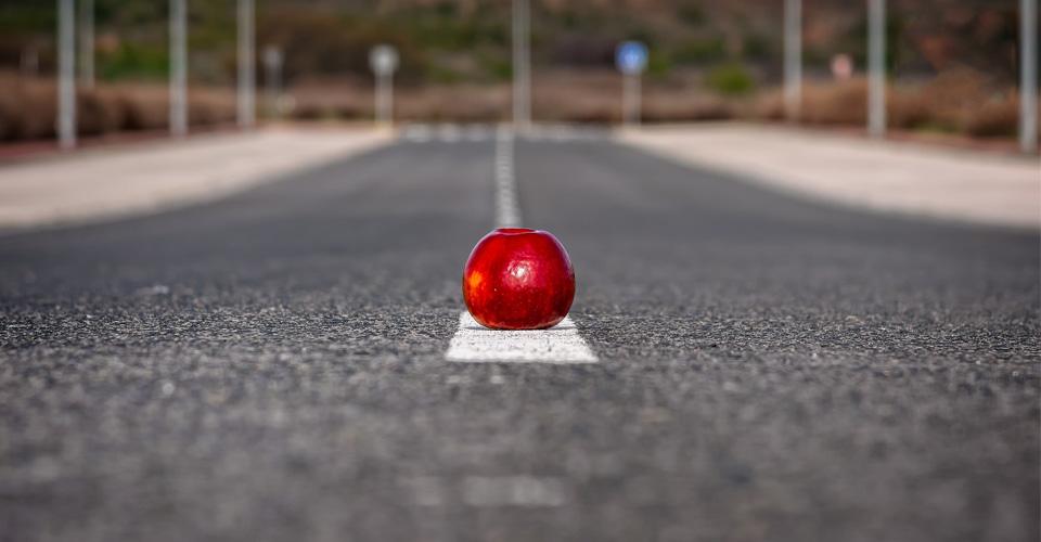 Ein glänzend roter Apfel liegt auf dem Mittelstreifen einer geraden Landstraße